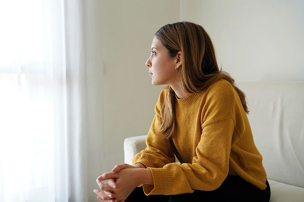 Eine junge Frau in einem gelben Pullover sitzt auf einer Couch und schaut nachdenklich aus einem Fenster mit weißen Vorhängen. Ihre Hände sind ineinander verschränkt, und ihr Gesichtsausdruck wirkt nachdenklich.