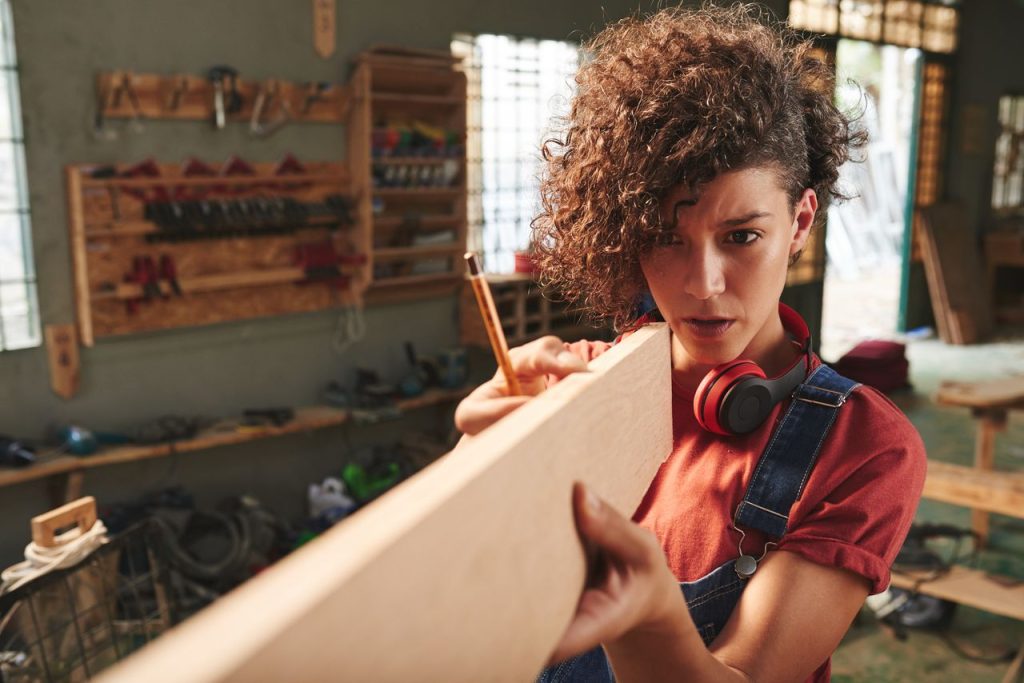 Eine junge Frau mit rotem T-Shirt steht in einer Tischler-Werkstatt und prüft mit gekonntem Blick die Geradlinigkeit einer zuvor gehobelten Holzlatte.