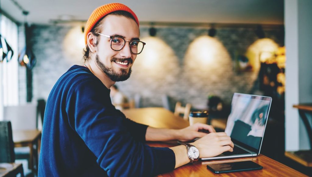 Ein Mann Anfang 20 mit Dreitagebart, Brille und Beanie-Mütze sitzt in einem Café und arbeitet am Laptop. Er blickt lächelnd in die Kamera.