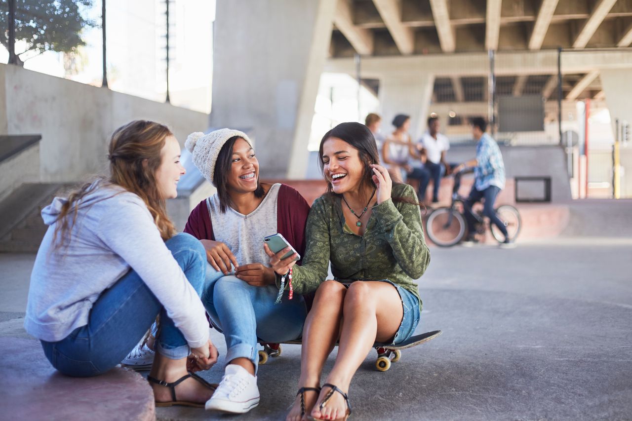 Drei junge Frauen unterschiedlicher Ethnien sitzen auf einem Skateboard in einem Skate-Park. Es herrscht sonniges Wetter. Im Hintergrund ist verschwommen eine weitere Gruppe junger Menschen zu sehen.