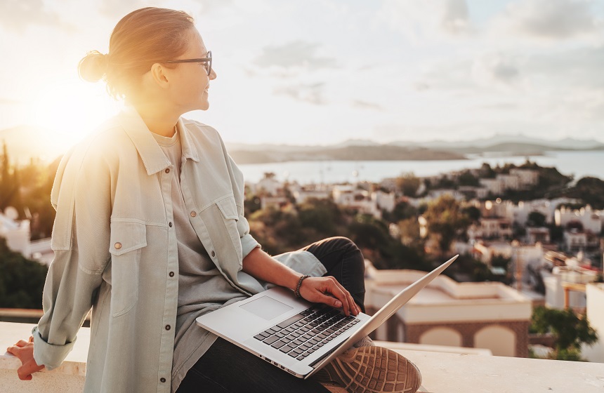 Eine junge Frau sitzt mit einem Laptop auf einer Mauer und sieht auf eine Stadt hinunter.