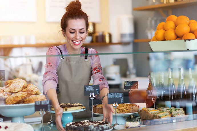 Eine Frau steht hinter dem Tresen eines Cafes und füllt die Auslage mit Kuchen