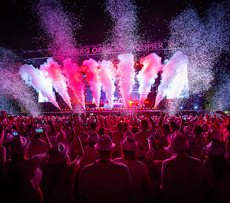 Eine große Menschenmenge in weißen Kleidern und Hüten jubelt bei einem nächtlichen Konzert. Von der Bühne, die mit rosa, roten und violetten Lichtern hell erleuchtet ist, schießen bunter Rauch und Konfetti.