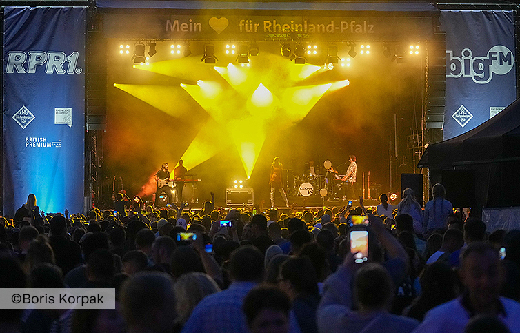 Eine große Menschenmenge schaut sich abends den Auftritt einer Band auf einer Freilichtbühne des Rheinland-Pfalz-Tags an. Fahnen an der Bühne zeigen die Logos der Radiosender big FM und RPR1.