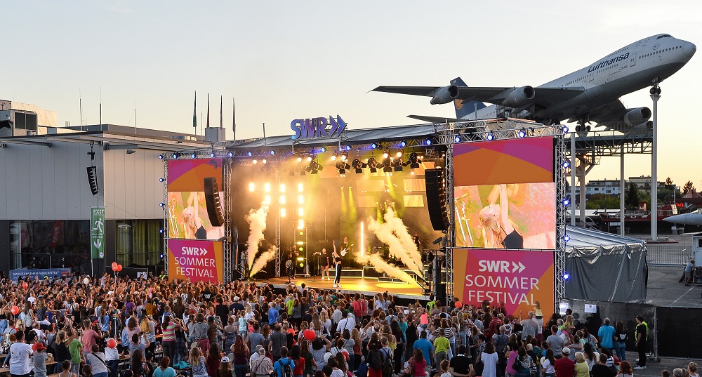 Eine große Menschenmenge versammelt sich vor einer Freilichtbühne beim SWR Sommerfestival mit hellen Scheinwerfern und Leinwänden. Im Hintergrund ist ein Lufthansa-Flugzeug über einem Gebäude in der Nähe des Festivalgeländes zu sehen.