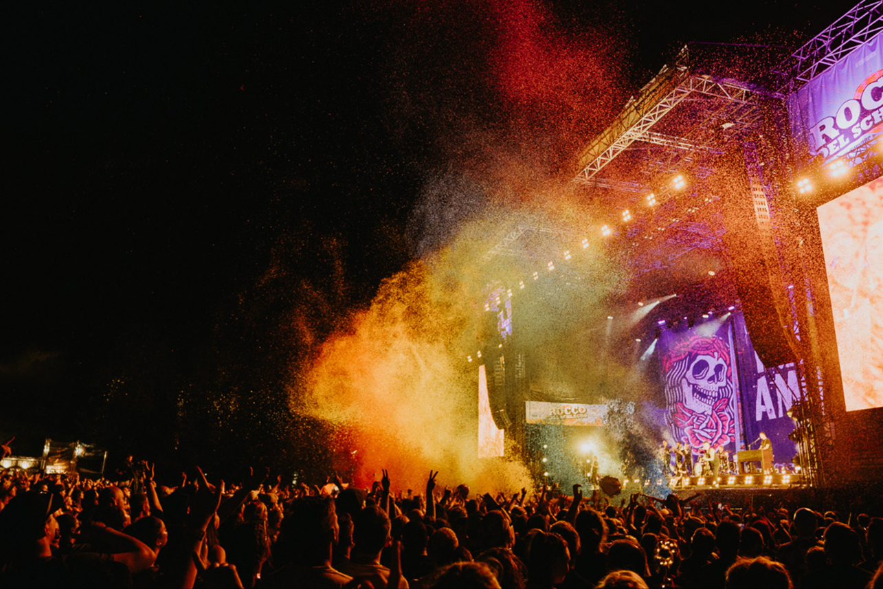 Eine große Menschenmenge genießt ein nächtliches Open-Air-Konzert. Buntes Pulver und Bühnenlichter erfüllen die Luft in der Nähe einer hell erleuchteten Bühne, die mit einem Totenkopfbanner dekoriert ist. Die Atmosphäre ist energiegeladen und festlich.