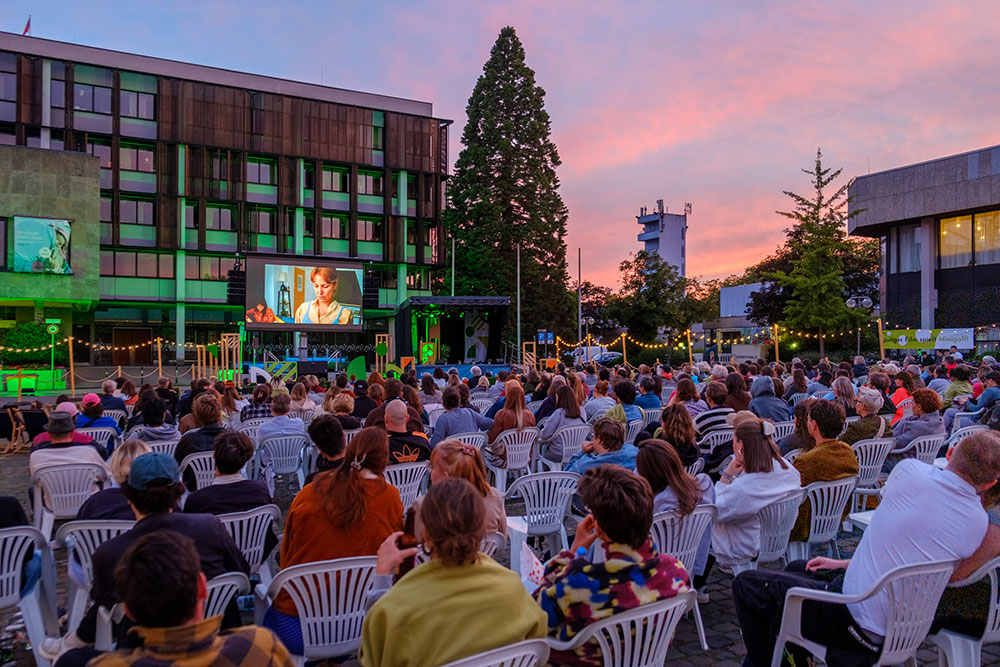 Eine große Menschenmenge sitzt draußen auf weißen Stühlen und schaut sich bei Sonnenuntergang einen Film auf einer großen Leinwand an. Lichterketten und Gebäude säumen den Platz und sorgen für eine festliche Atmosphäre.