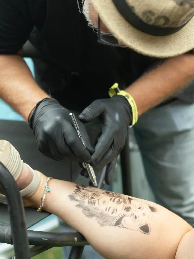 Eine Person mit schwarzen Handschuhen und Hut tätowiert einen detailreichen Wolf auf einen Unterschenkel. Sandale und Fußkettchen sind sichtbar.