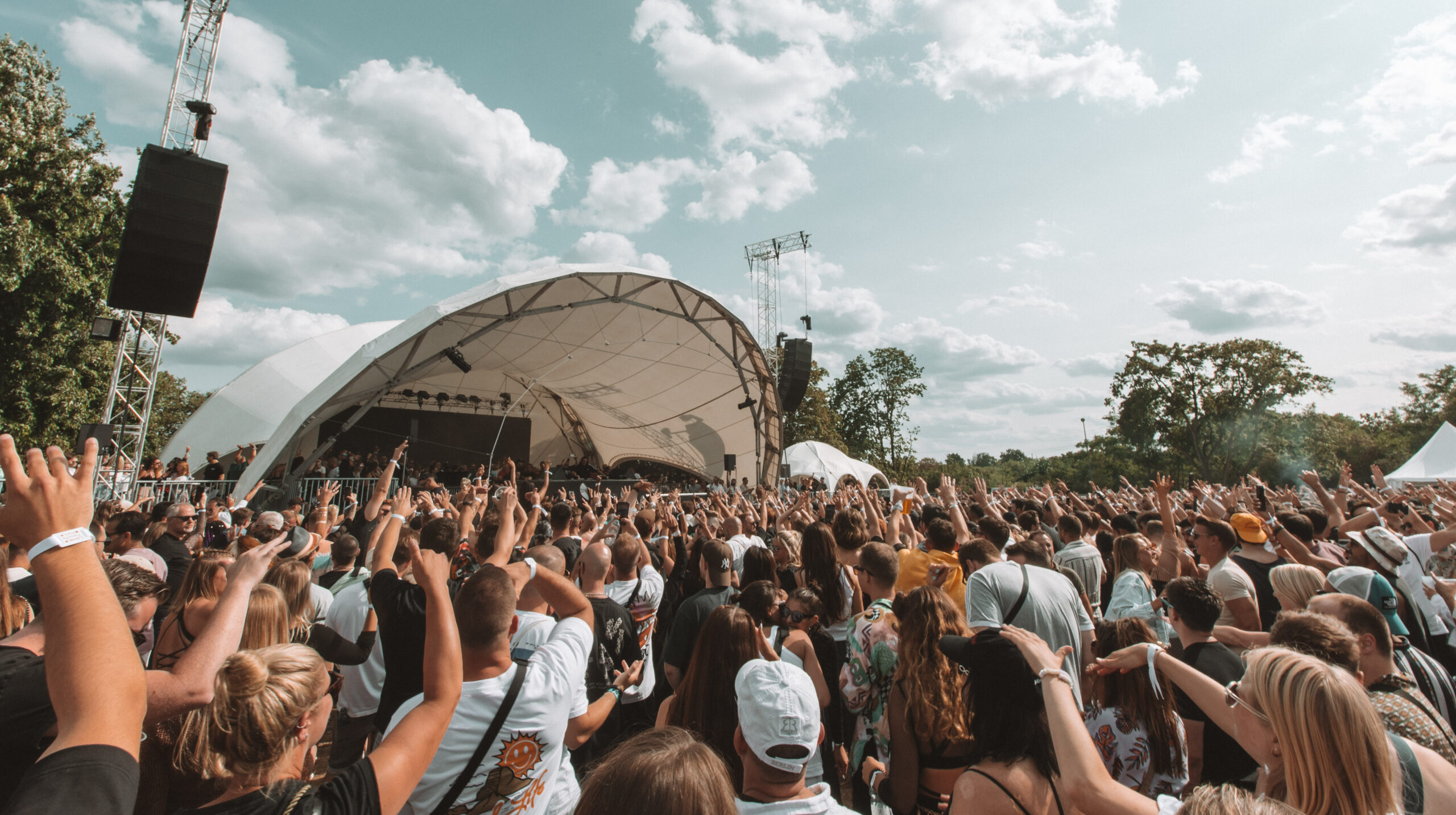 Eine große Menschenmenge mit erhobenen Händen steht vor einer Freilichtbühne mit weißem Baldachin und genießt an einem sonnigen Tag mit vereinzelten Wolken eine Live-Musik-Darbietung. Im Hintergrund sind Bäume und Veranstaltungsstrukturen zu sehen.