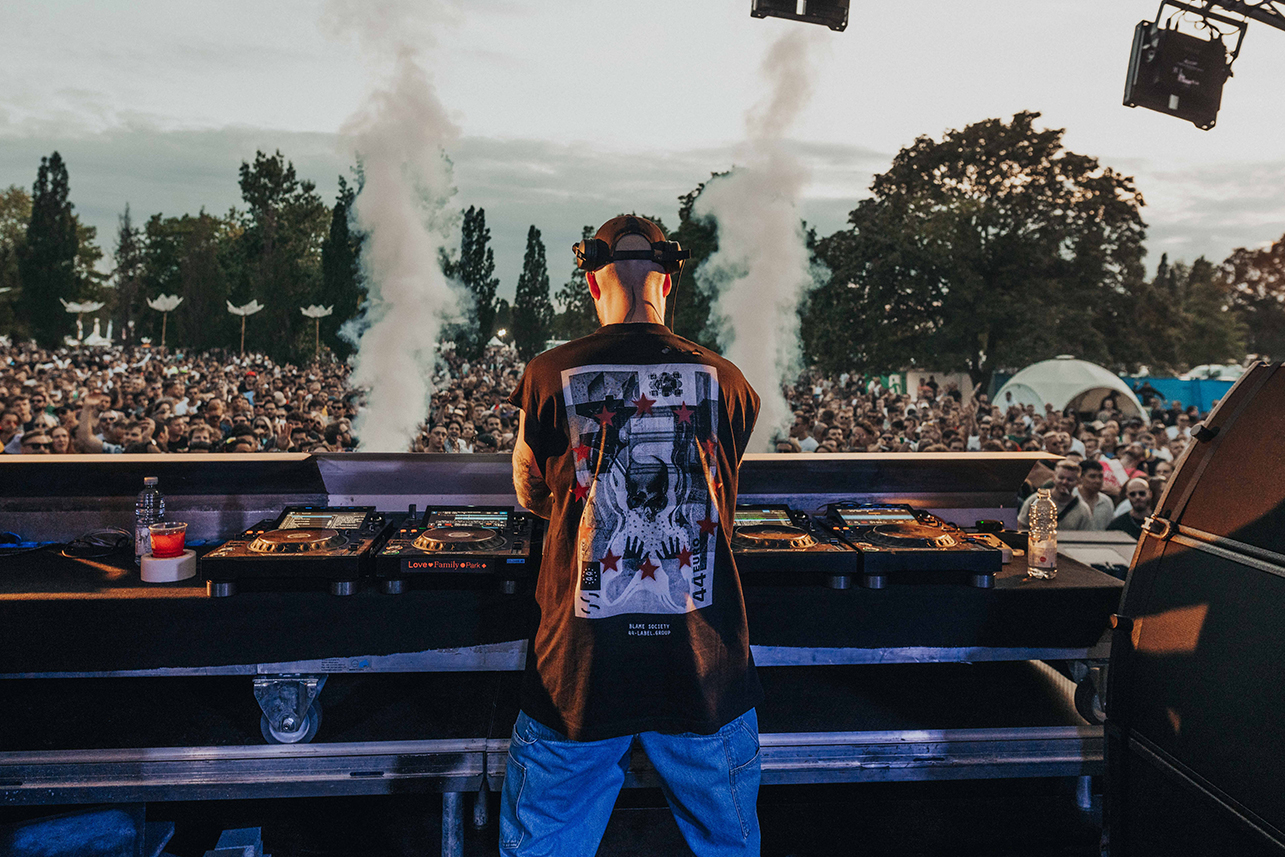 Ein DJ steht an einem Mischpult auf einer Freilichtbühne vor einer großen Menschenmenge bei einem Musikfestival. Hinter ihm stoßen Nebelmaschinen Rauchschwaden aus. Im Hintergrund sind Bäume und Zelte unter einem bewölkten Himmel zu sehen.