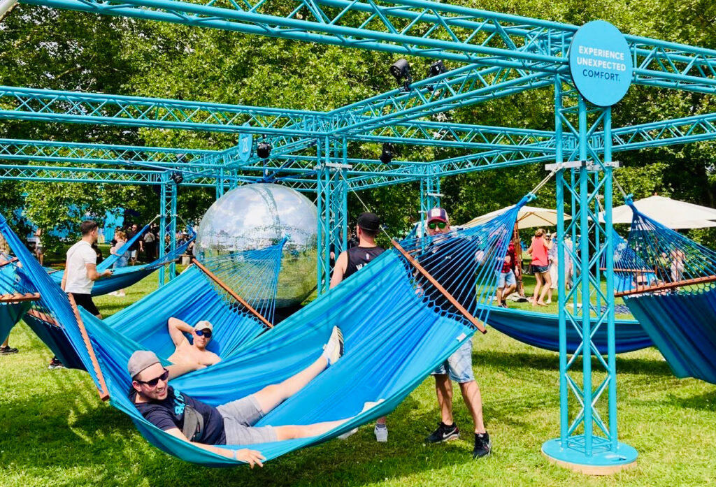 Menschen entspannen in blauen Hängematten unter einer Metallkonstruktion in einem Park. In der Mitte befindet sich eine große Spiegelkugel. Im Hintergrund sind Bäume und andere Menschen zu sehen. Auf einem Schild steht „Erleben Sie unvollkommenen Komfort“.
