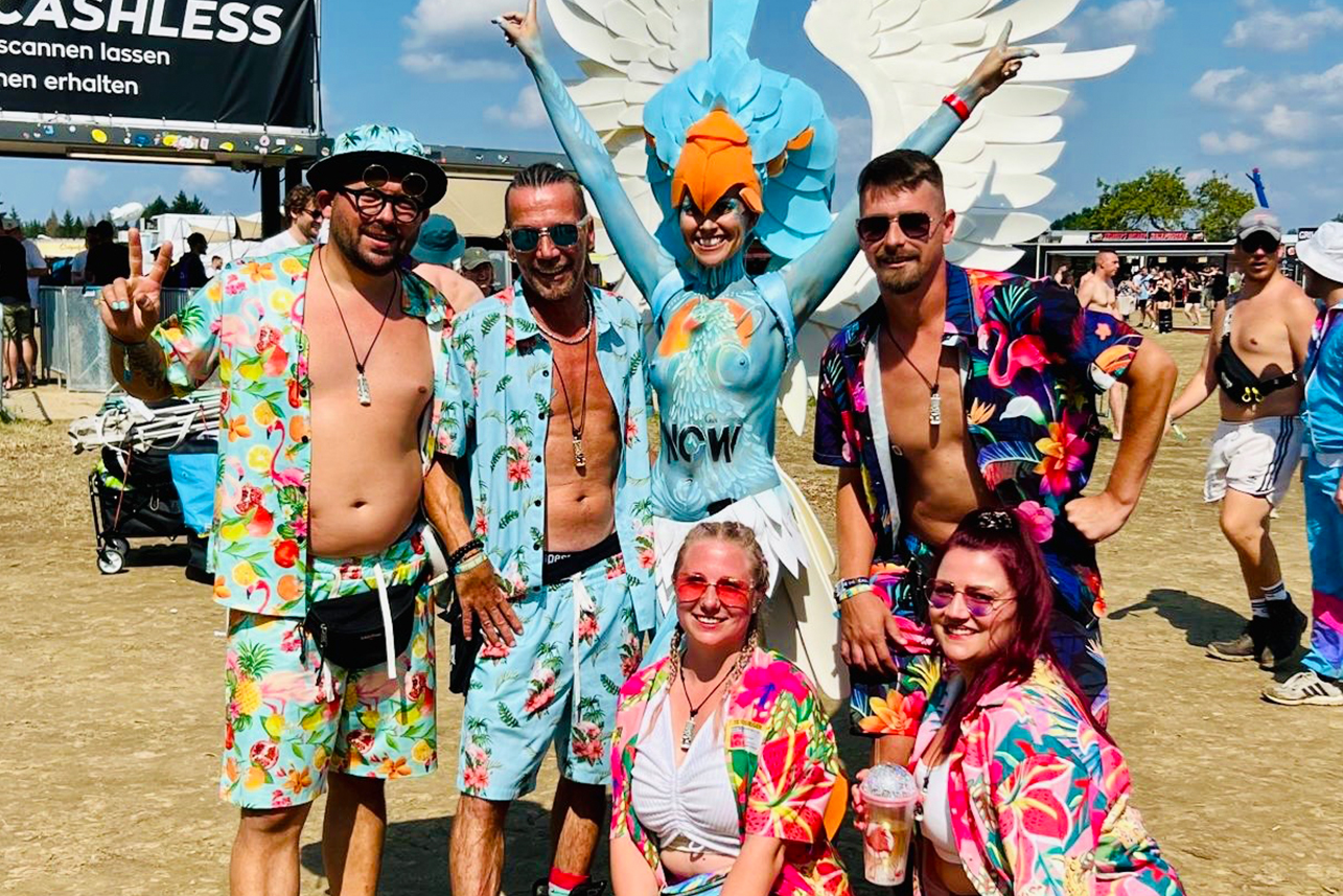 Eine Gruppe von fünf Personen in farbenfrohen Blumenkostümen posiert lächelnd auf einem Festival. Eine Künstlerin mit Bodypainting und großen weißen Flügeln steht mit erhobenen Armen da. Im Hintergrund sind Festivalzelte und Besucher zu sehen.