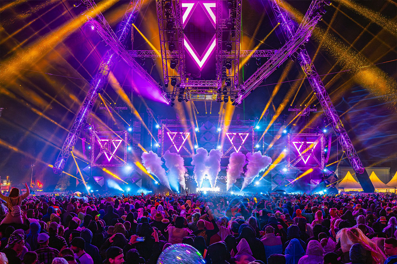 Ein pulsierendes Musikfestival am Abend mit einer großen Menschenmenge, bunten Lichtern, Raucheffekten und geometrischen Lichtspielen auf der Bühne unter einem Metallgerüst. Das Publikum blickt zur Bühne und genießt die energiegeladene Atmosphäre.