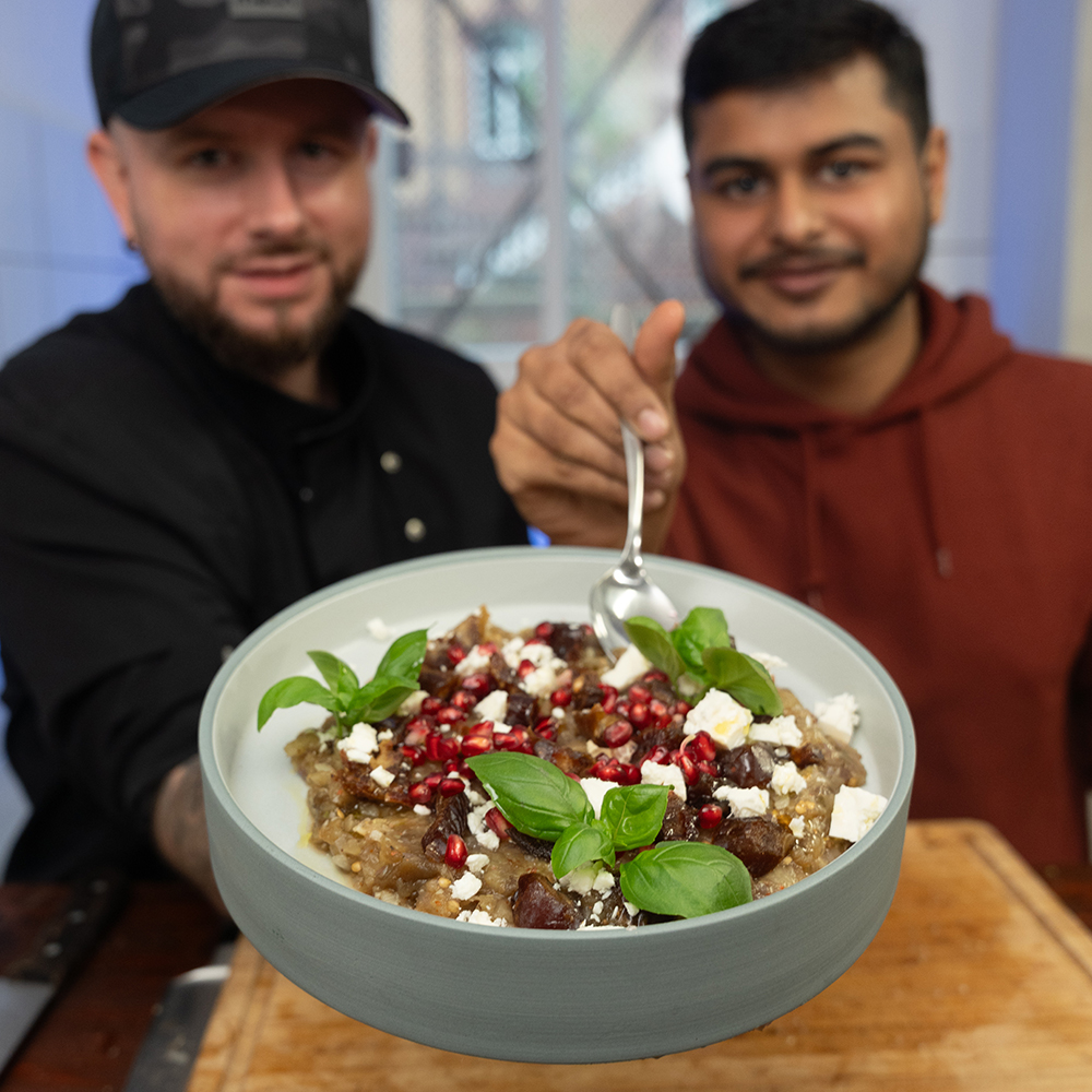 Zwei Männer präsentieren eine Schüssel mit Feta, Granatapfelkernen und Basilikum. Einer hält einen Löffel voll und streckt ihn in Richtung Kamera. Das Gericht scheint auf einer Holzplatte in einer Küchenumgebung zu stehen.
