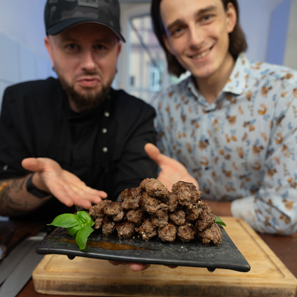 Zwei Männer, einer in schwarzer Kochkleidung und -mütze, der andere in einem gemusterten Hemd, präsentieren in einer Küchenumgebung eine Platte mit gegrillten Fleischstücken, garniert mit Basilikum, und lächeln in die Kamera.