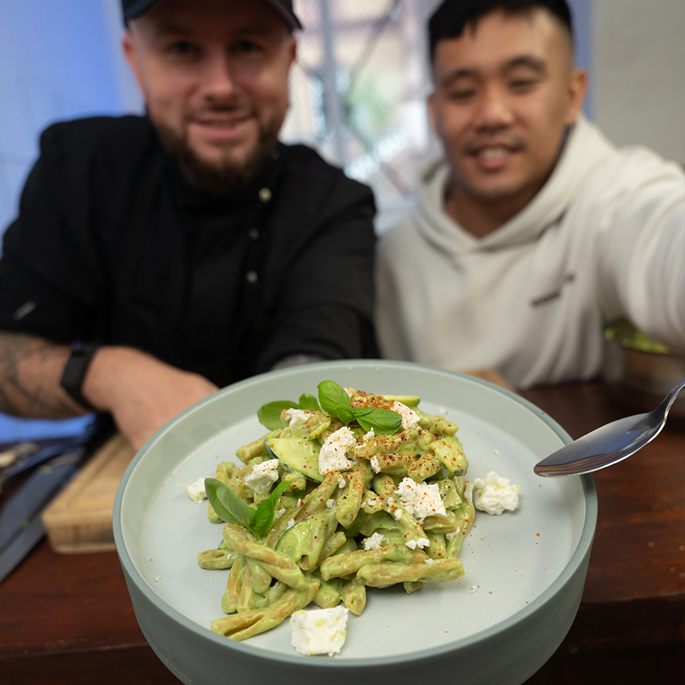 Zwei lächelnde Menschen stehen hinter einem Teller Pasta mit grüner Sauce, frischen Kräutern und geriebenem Käse. Das Gericht ist im Vordergrund scharf dargestellt, während die Menschen im Hintergrund leicht unscharf sind.