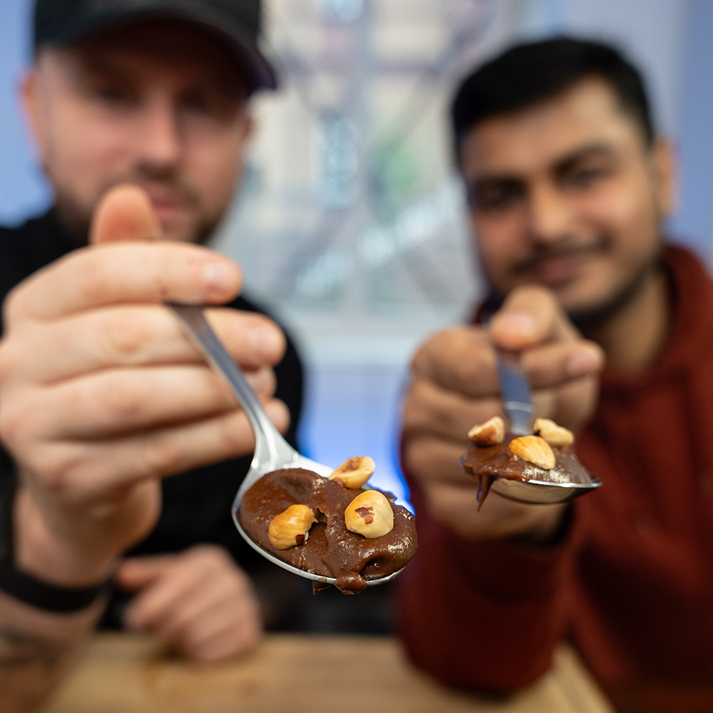 Zwei Personen halten Löffel mit Schokoladenaufstrich und Haselnüssen in die Kamera. Ihre Gesichter sind im Hintergrund unscharf. Das Bild hebt das Dessert auf den Löffeln hervor.