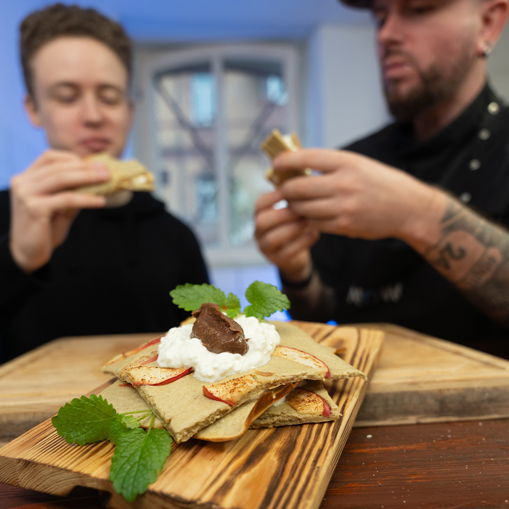 Zwei Personen sitzen an einem Tisch und halten Crêpes in den Händen. Im Vordergrund sind auf einem Holzbrett gefaltete Crêpes mit Schlagsahne, Schokolade und Minzblättern zu sehen. Der Hintergrund ist unscharf.