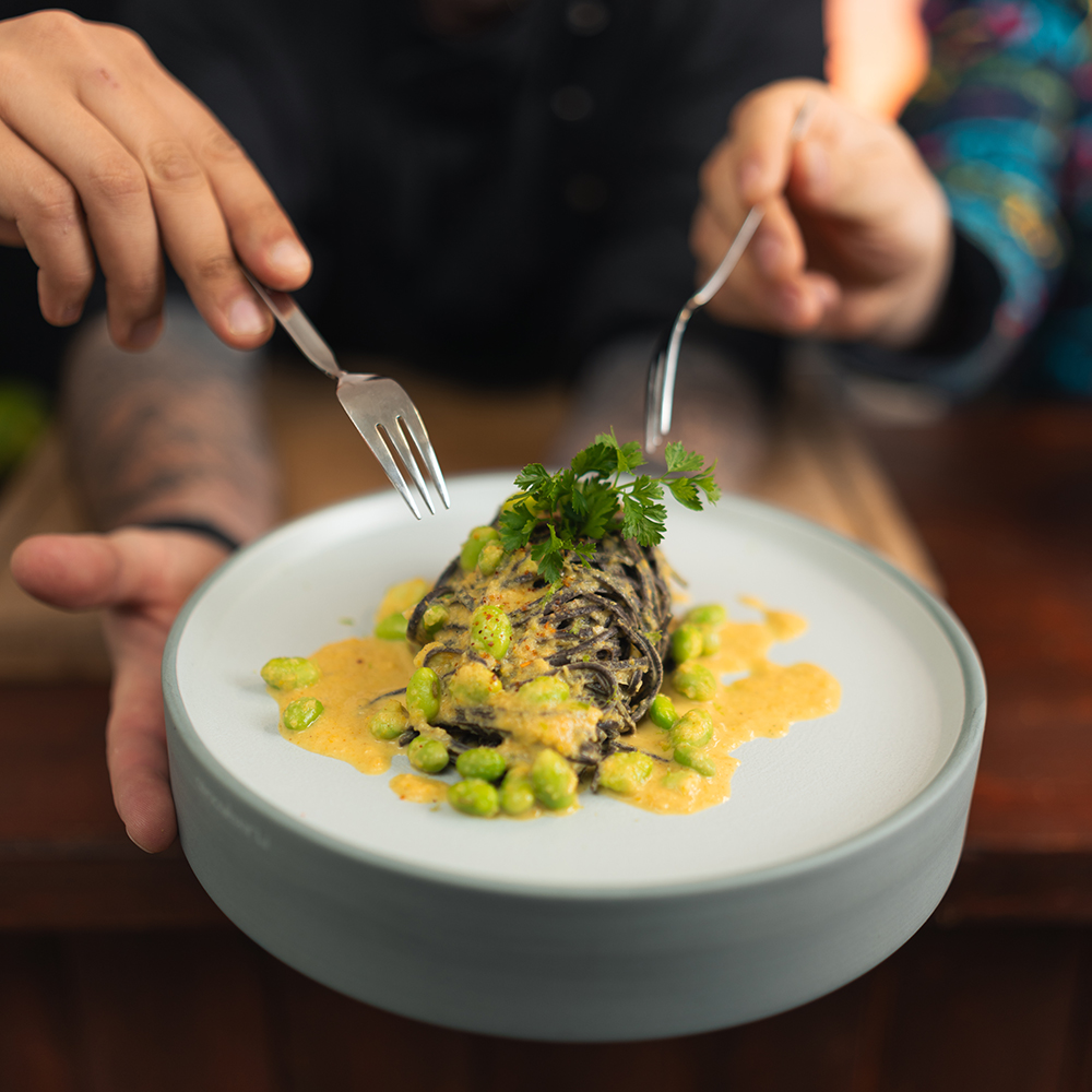 Eine Person hält einen Teller mit einem Gourmetgericht mit frischen Kräutern und cremiger Soße, garniert mit grünen Bohnen. Zwei Hände mit Gabeln strecken sich, um das Essen zu teilen.