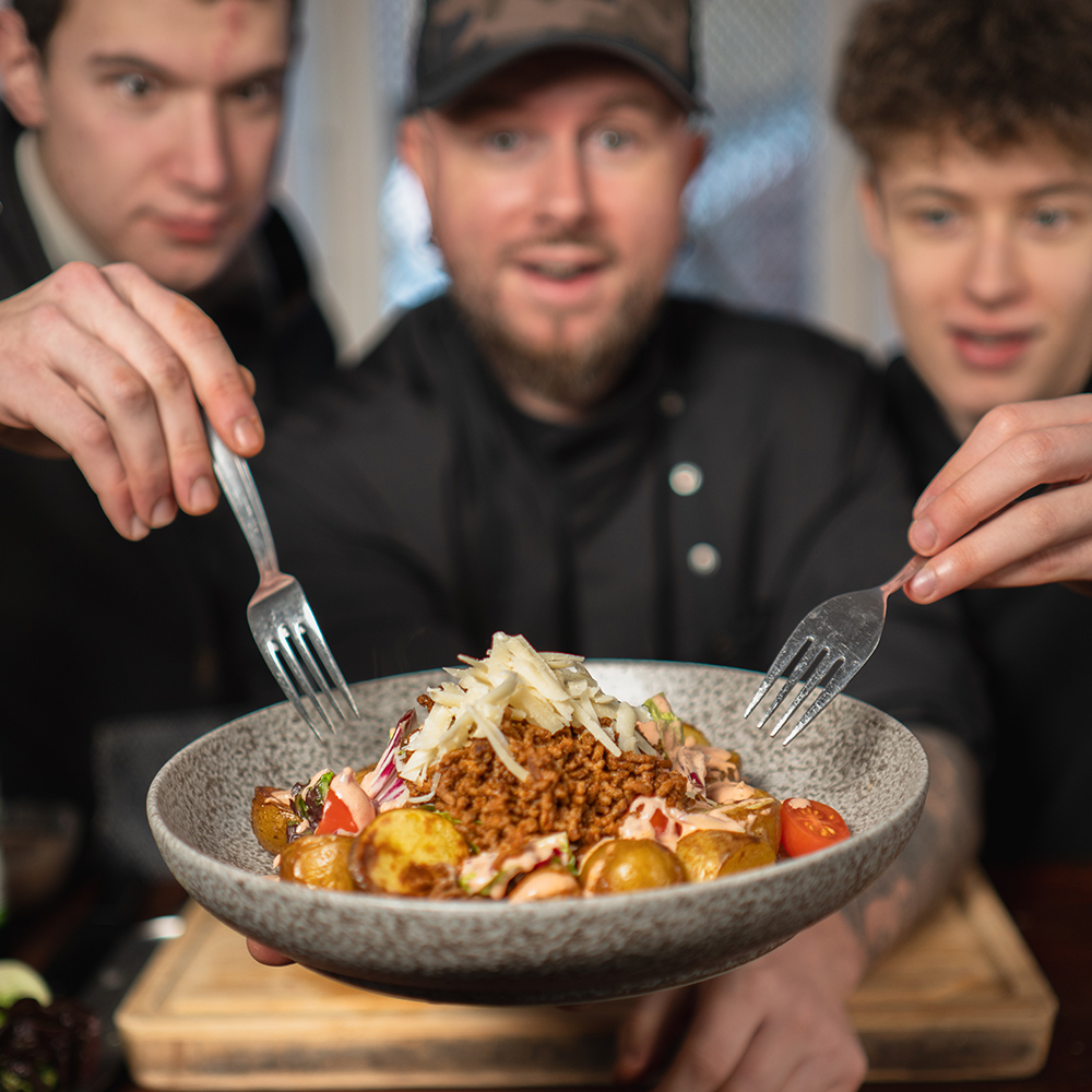 Drei Personen halten aufgeregt Gabeln in Richtung einer Schüssel mit gebratenen Kartoffeln, geraspeltem Gemüse und geriebenem Parmesan, die auf einem grauen Teller präsentiert wird. Der Hintergrund ist leicht unscharf und konzentriert sich auf das Essen und ihre Reaktionen.