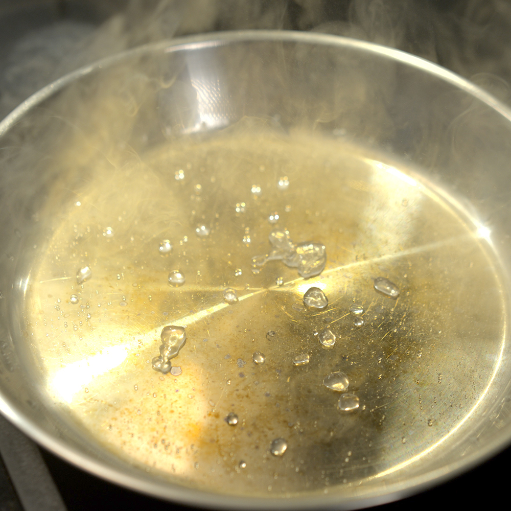 Nahaufnahme einer heißen, leeren Metallbratpfanne, auf deren Oberfläche Wassertropfen brutzeln und dampfen. Die Hitze lässt sichtbaren Dampf aus der Pfanne aufsteigen.