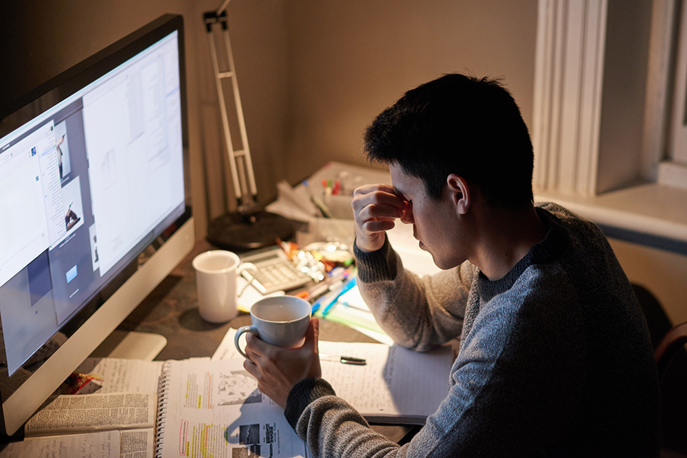 Eine Person sitzt an einem unordentlichen Schreibtisch mit aufgeschlagenem Notebook und Computer, hält eine Tasse in der Hand und reibt sich die Stirn, wirkt gestresst oder müde, während sie studiert oder bis spät in die Nacht arbeitet.