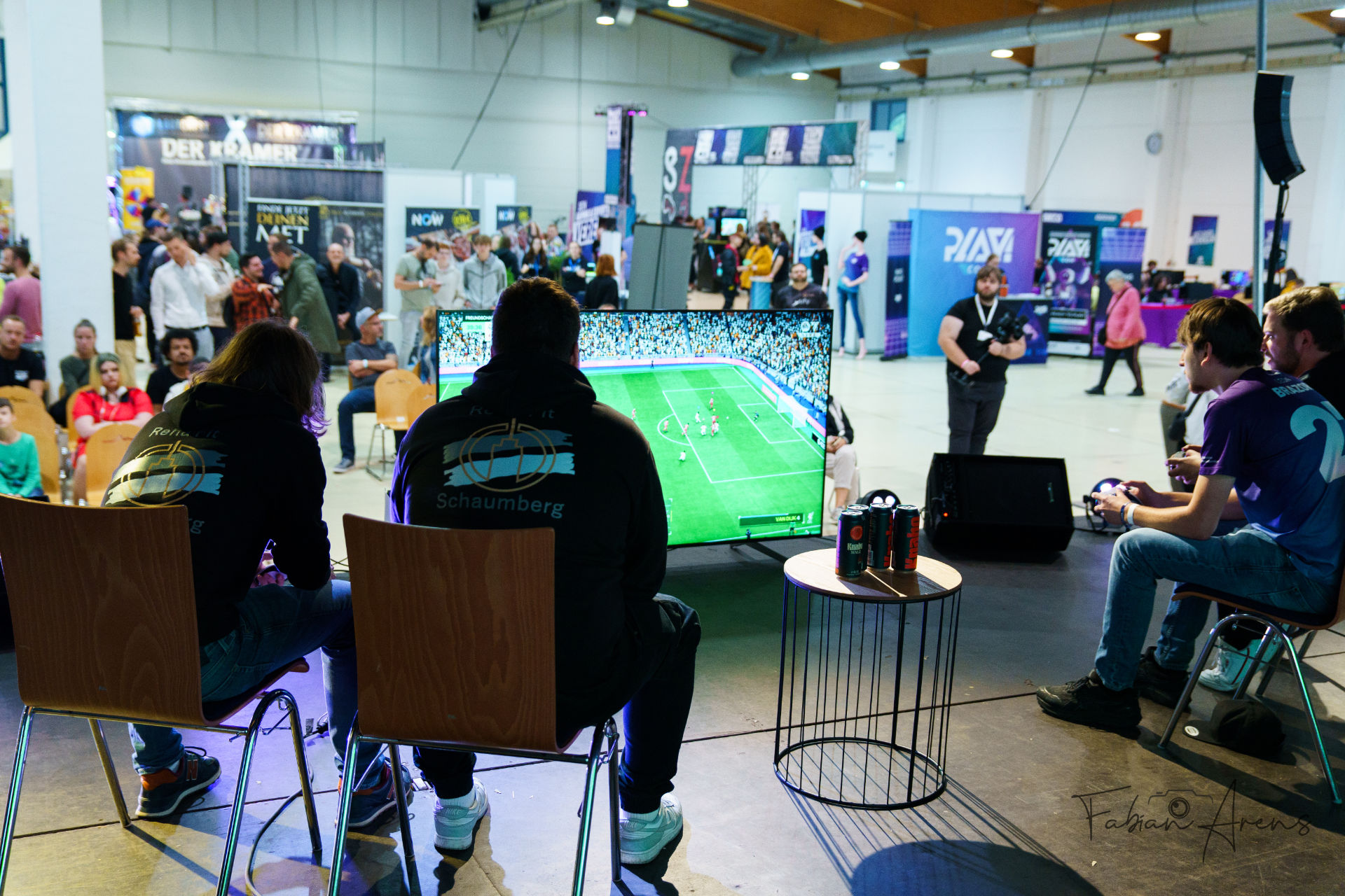 Zwei Personen sitzen auf Stühlen auf einer Bühne und spielen ein Fußball-Videospiel auf einem großen Bildschirm, während eine Menschenmenge bei einem Gaming-Event zusieht. Im Hintergrund sind mehrere Stände und Banner zu sehen.