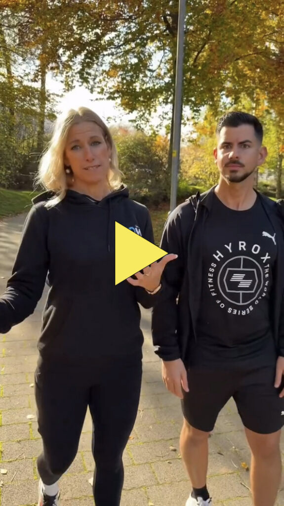 Zwei Personen stehen an einem sonnigen Tag im Freien und tragen sportliche Kleidung. Die Frau auf der linken Seite gestikuliert mit ihren Händen, während der Mann auf der rechten Seite mit den Armen an den Seiten steht. Das Bild ist mit einer gelben Play-Taste überlagert.