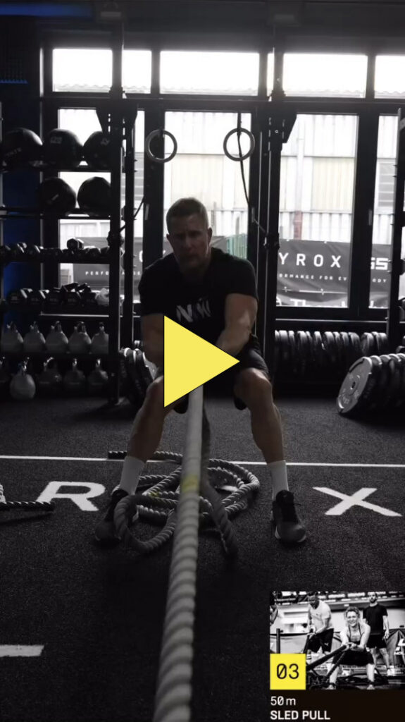 Ein Mann in einem Fitnessstudio zieht ein dickes Seil zu sich heran, während er breitbeinig und mit ausgestreckten Armen sitzt. Im Hintergrund sind Fitnessgeräte wie Kettlebells und Gewichte zu sehen. Eine gelbe Play-Taste überlagert das Bild.