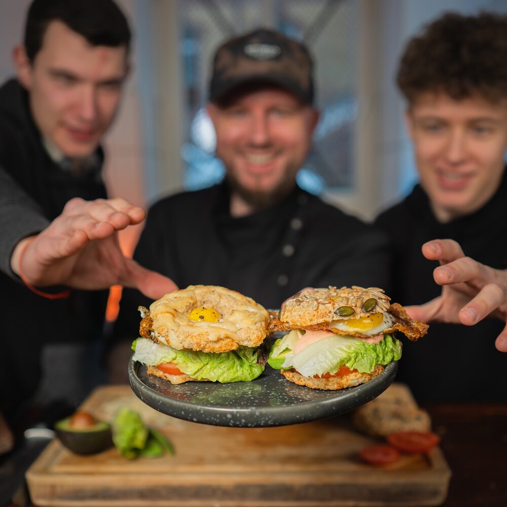 Drei lächelnde Männer präsentieren vor einem rustikalen Küchenhintergrund zwei Gourmet-Bagel mit verschiedenen Belägen, die auf einem Metallspatel liegen.