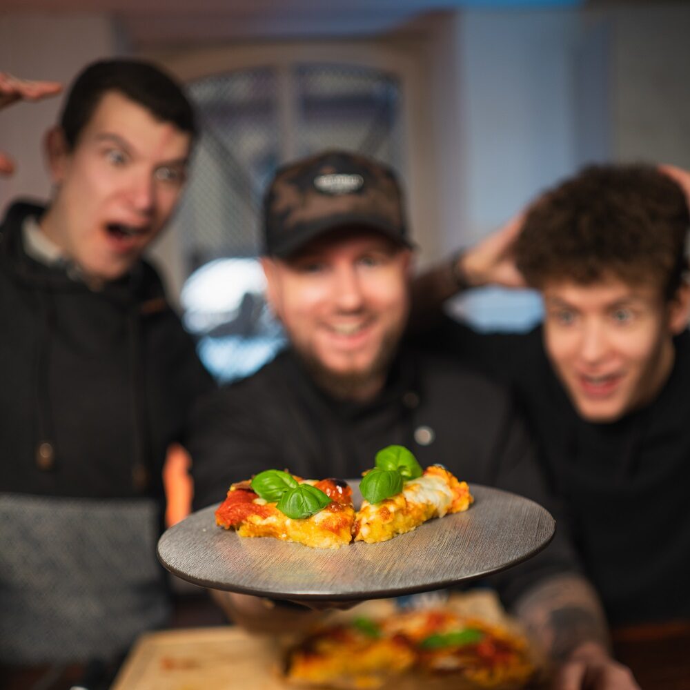 Drei Männer lächeln und sehen aufgeregt aus, als sie eine mit Basilikum belegte Pizzascheibe im Vordergrund präsentieren, während der Rest der Pizza auf einem hölzernen Schneidebrett hinter ihnen liegt. Der Fokus liegt auf der Pizzascheibe, die aus Kartoffeln hergestellt wurde