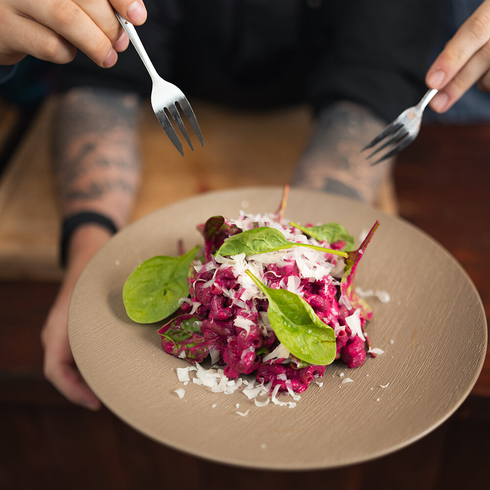 Eine Person präsentiert einen beigen Teller mit Erbsen Pasta mit Rote Bete Feta Sauce der mit Blattspinat und geriebenem Parmesan belegt ist, während zwei Hände mit Gabeln nach dem Essen greifen.
