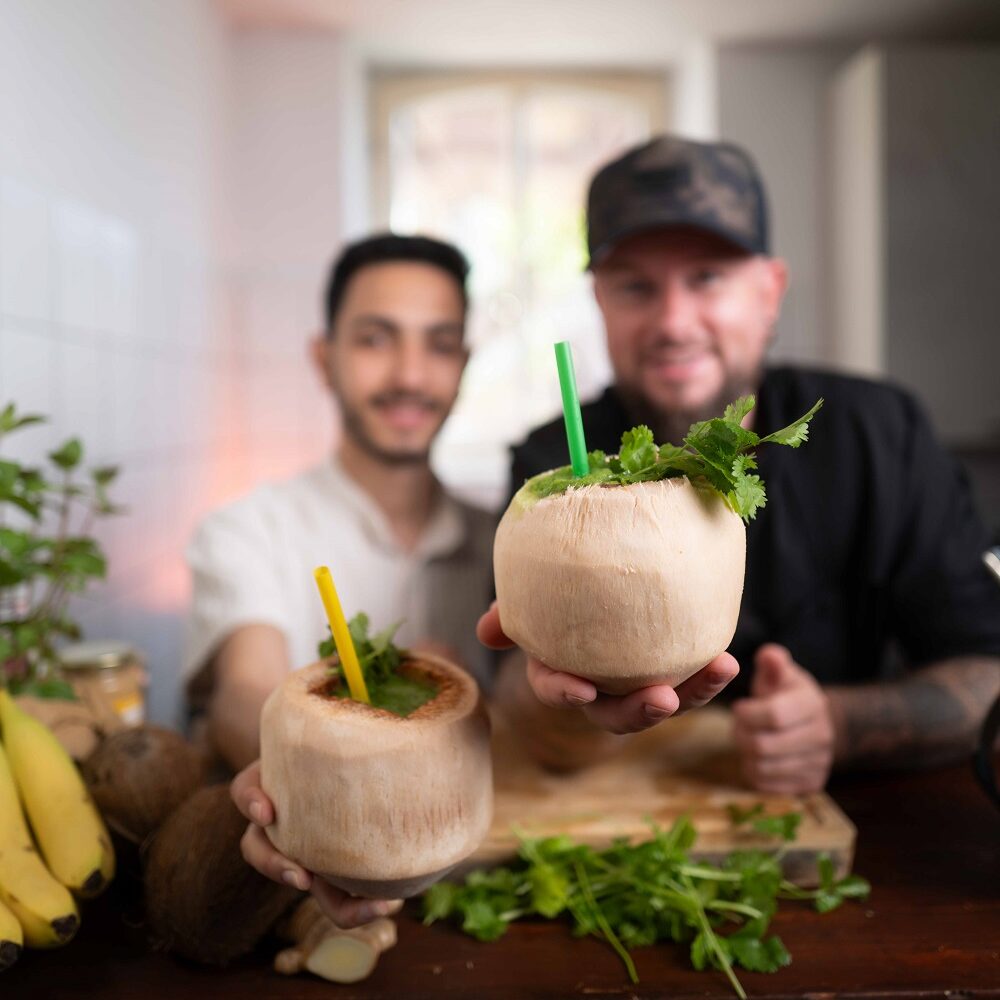 Zwei Männer in einer Küche halten frische Kokosnüsse mit bunten Strohhalmen und mit Kräutern garniert in die Höhe. Auf der Theke vor ihnen liegen Bananen und frisches Grün. Der Hintergrund ist sanft verschwommen.