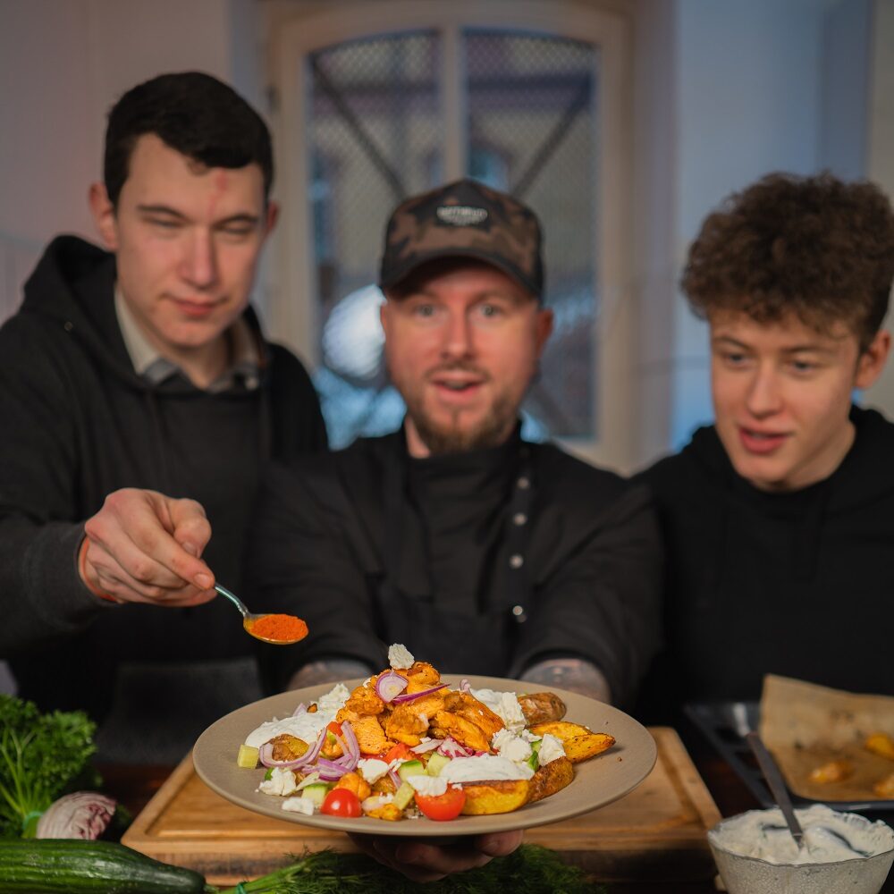 Drei Männer stehen zusammen in einer Küche. Der vordere Mann hält einen Teller mit Salat, Gemüse und Käse in der Hand, während ein anderer einen Löffel mit roter Soße reicht. Sie schauen alle auf das Essen, wirken glücklich und beschäftigt.