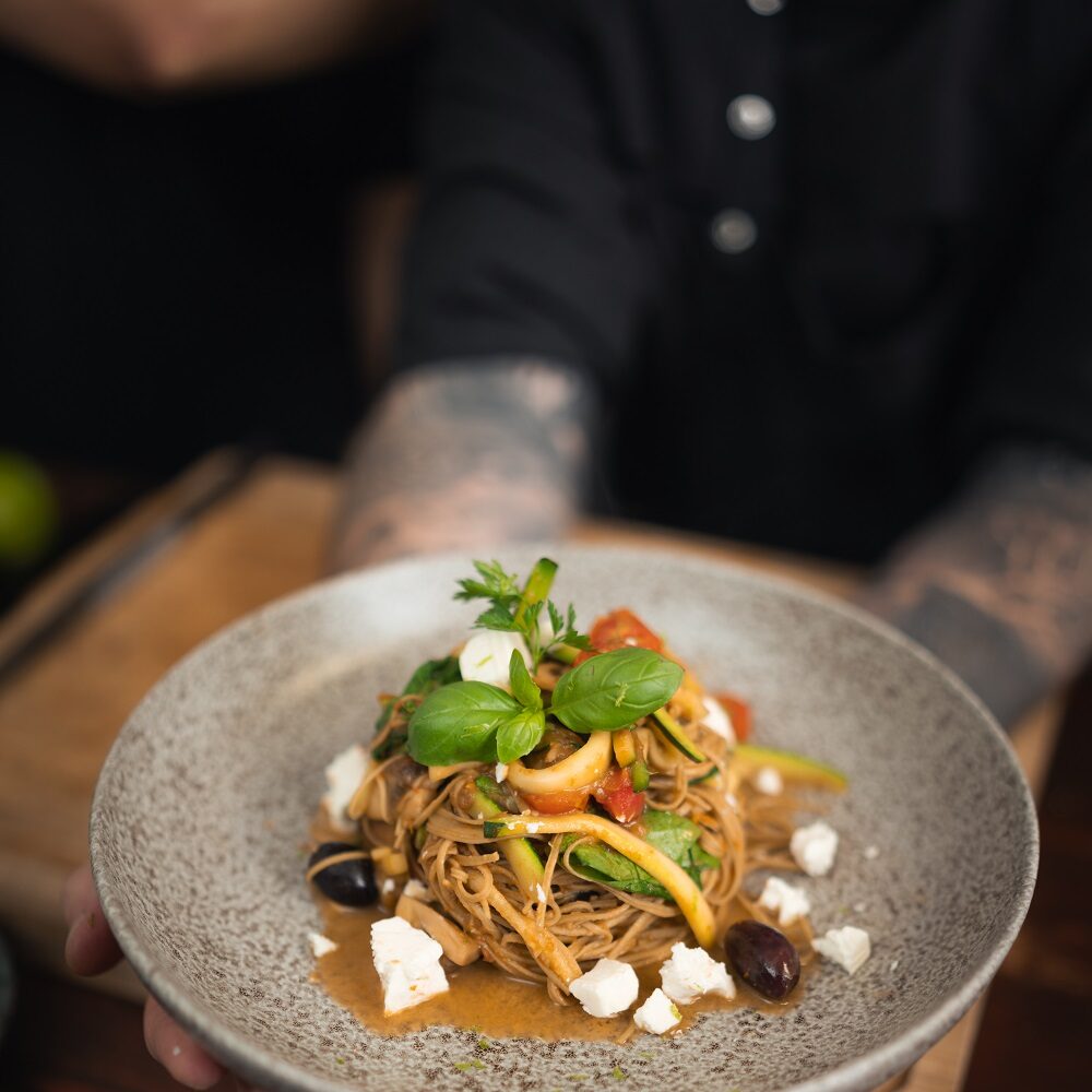 Eine Person mit tätowierten Armen hält eine grau gesprenkelte Schüssel mit Nudeln, garniert mit frischen Kräutern, Oliven und zerbröckeltem Käse, über einen dunklen Holztisch.