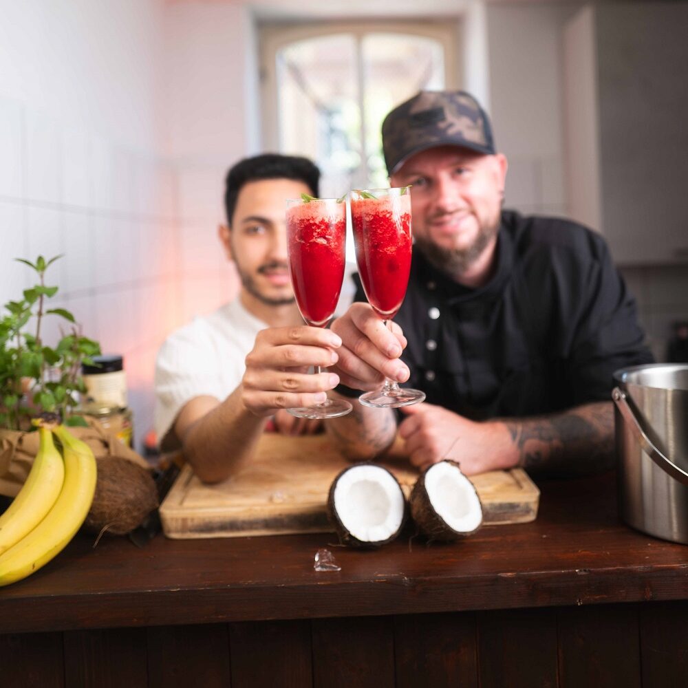 Zwei Männer lächeln und halten Gläser mit einem roten Getränk in einer Küche hoch, auf der Theke vor ihnen liegen Früchte wie Bananen und eine aufgeschnittene Kokosnuss.