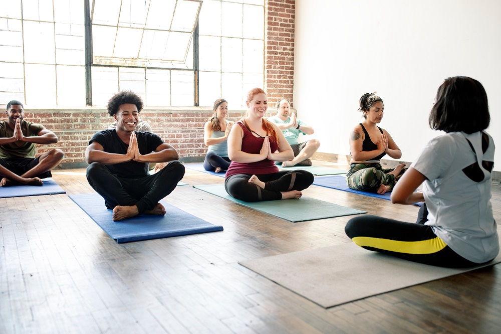 Eine Gruppe von Menschen sitzt im Schneidersitz auf Yogamatten in einem hellen Studio und folgt einem Lehrer an der Spitze. Sie lächeln und haben ihre Hände in einer Gebetshaltung zusammen.