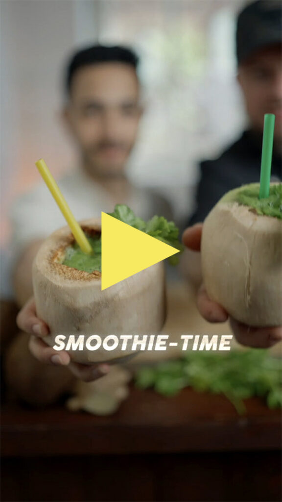 Zwei Personen halten Kokosnüsse mit grünem Smoothie und Strohhalmen in die Höhe. Der Text SMOOTHIE-TIME und eine gelbe Play-Taste erscheinen über dem Bild. Der Hintergrund ist unscharf.
