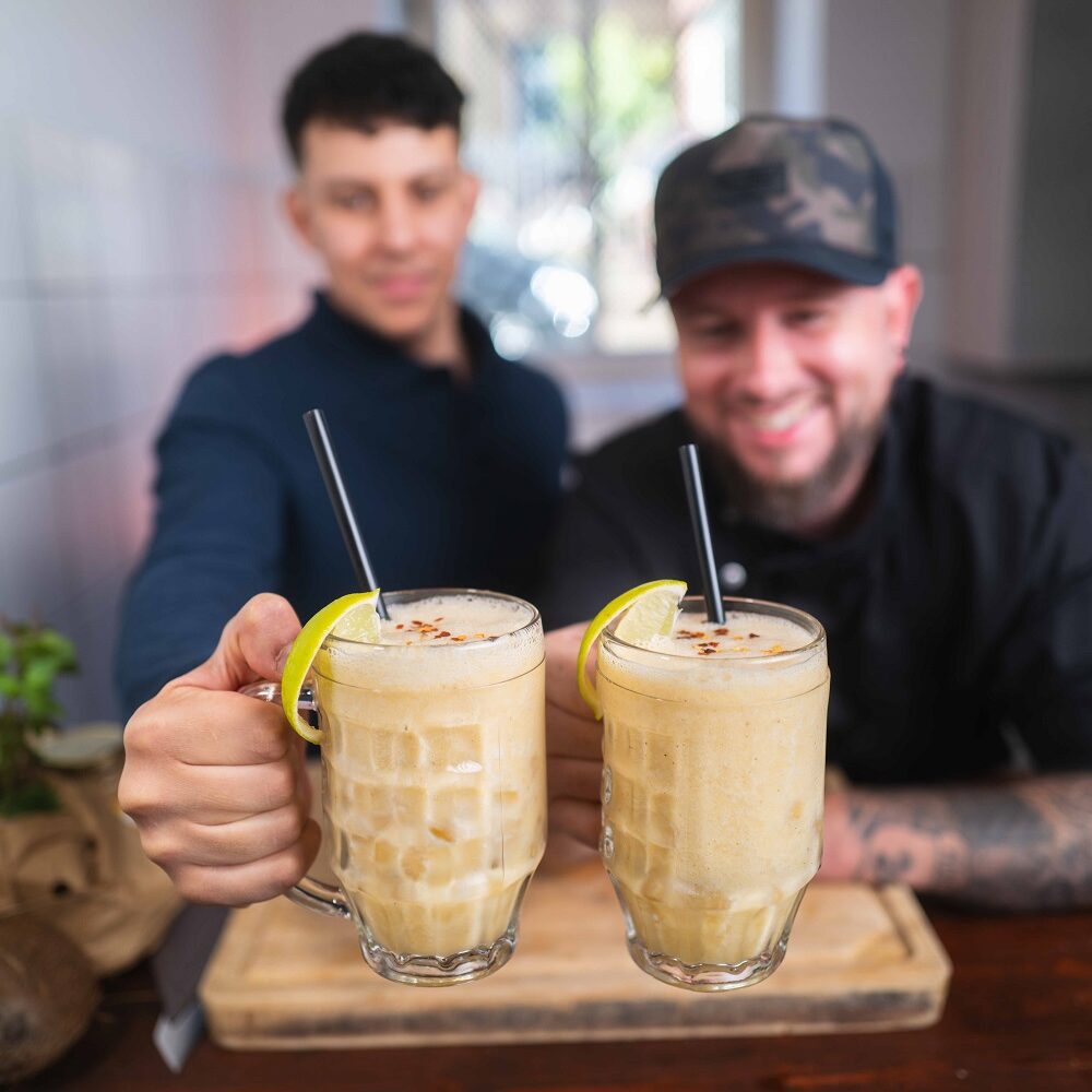 Zwei lächelnde Männer halten einen gelben Smoothie in Glasbechern, garniert mit Limettenscheiben und Strohhalmen, in einer gemütlichen, sonnenbeschienenen Küche in die Höhe. Der Fokus liegt auf den Getränken im Vordergrund.