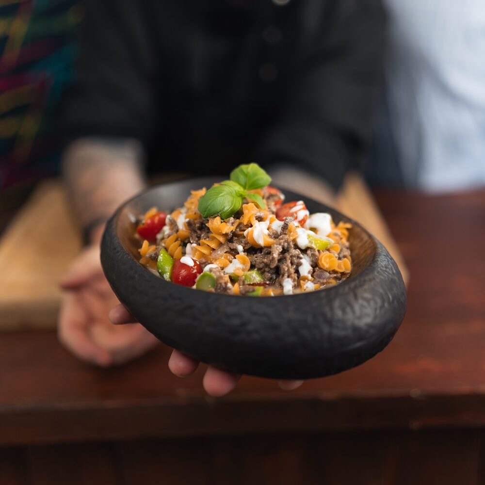 Ein lächelnder Koch präsentiert eine schwarze Schüssel mit einem bunten Gericht aus Nudeln, Hackfleisch, Kirschtomaten, Paprika, Käse und Basilikum, während im Hintergrund zwei Personen stehen.