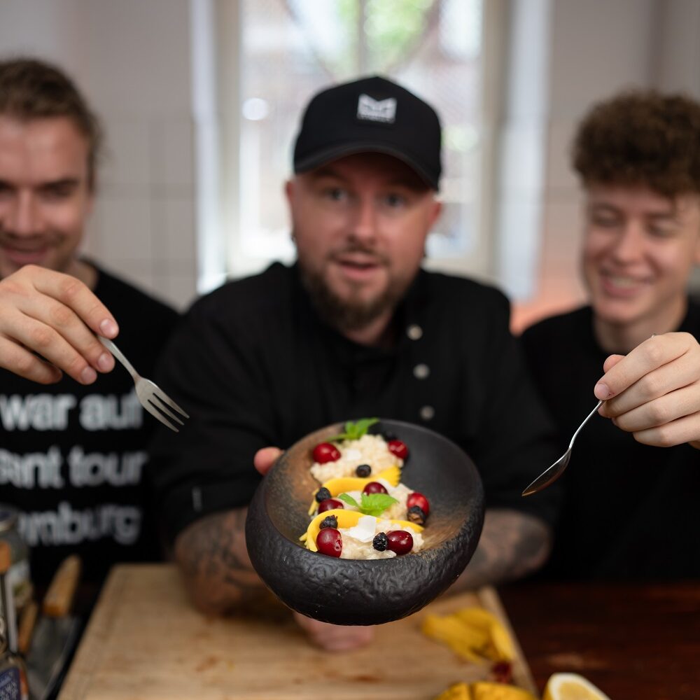 Drei Personen sitzen an einem Holztisch. Die Person in der Mitte, die eine schwarzen Mütze und eine schwarze Jacke trägt, hält eine Schüssel mit buntem Essen, das mit Granatapfelkernen bestückt ist. Die anderen greifen mit Gabeln hinein und lächeln. Auf dem Tisch liegen Mango und Zitrone in Scheiben geschnitten.