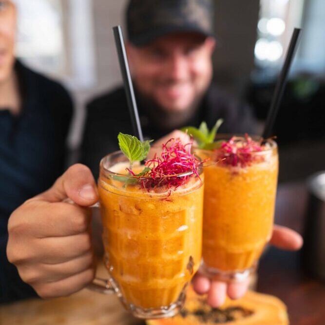 Zwei Personen halten cremig-orangefarbene Smoothies in Glasbechern, garniert mit Kräutern und roten Sprossen, mit schwarzen Strohhalmen hoch. Eine Papayahälfte liegt auf dem Holztisch vor ihnen. Beide lächeln im Hintergrund.