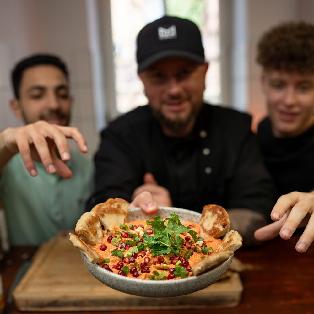 Drei Personen greifen nach einer Schale Hummus mit Kräutern und Gewürzen, umgeben von Pita-Scheiben, und in der Mitte steht ein Koch, der das Gericht in einer Küchenzeile hält.