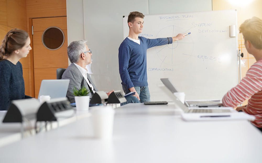 Ein junger Mann steht und zeigt auf ein Whiteboard mit Notizen, während drei Prüfer mit Laptops an einem Konferenztisch sitzen.