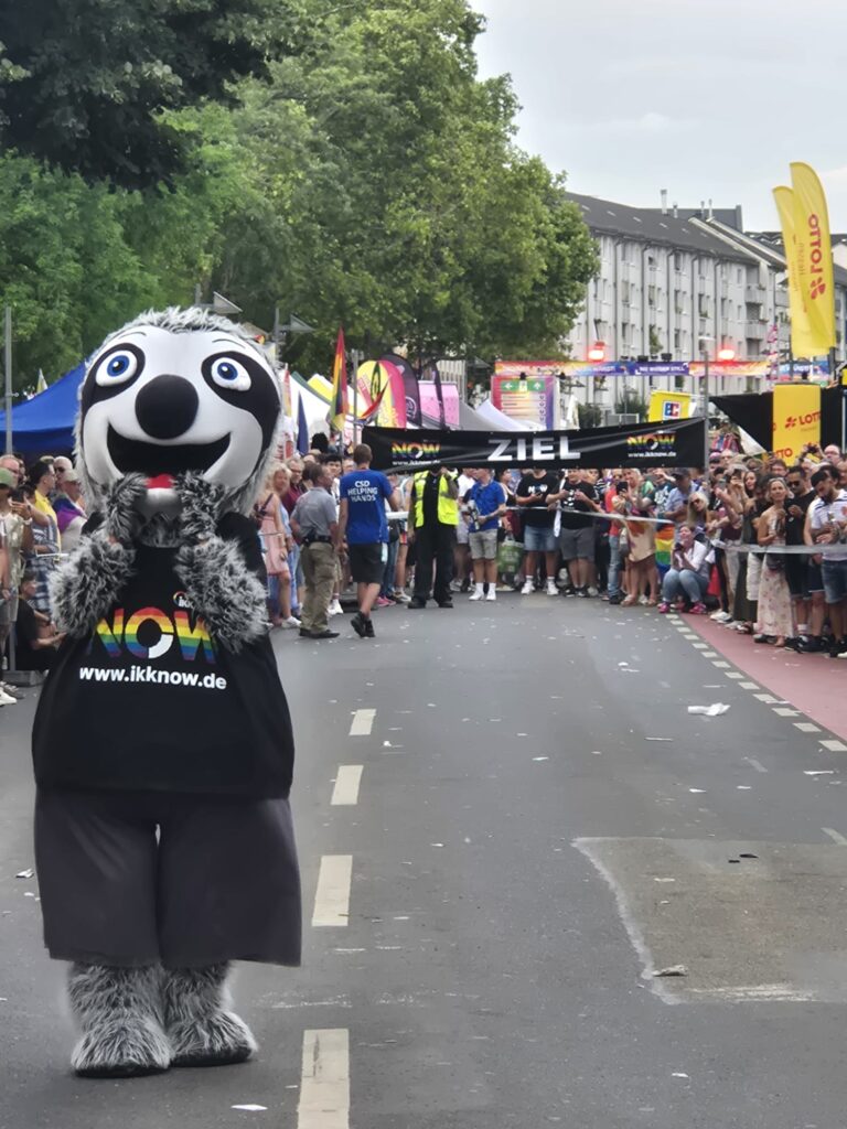Eine Person in einem fröhlichen Faultier-Maskottchen-Kostüm steht während einer öffentlichen Veranstaltung auf einer Straße. Die Menschenmenge versammelt sich hinter einem Zielbanner mit der Aufschrift ZIEL und bunten Fahnen und Bannern an den Seiten.