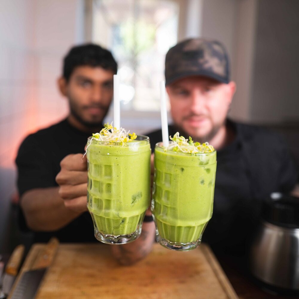 Zwei Männer halten Gläser mit grünem Smoothie mit Strohhalmen und gelber Blumengarnitur in der Hand und stehen in einer Küche mit einer hölzernen Arbeitsplatte im Vordergrund.