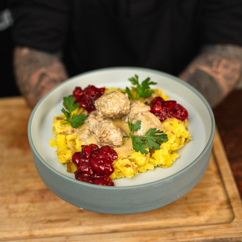 Eine Person hält eine Schüssel mit schwedischen Fleischbällchen in Sahnesoße, serviert mit Kartoffelpüree, Preiselbeersoße und garniert mit Petersilie. Der Hintergrund zeigt eine Holzfläche und die Arme der Person.