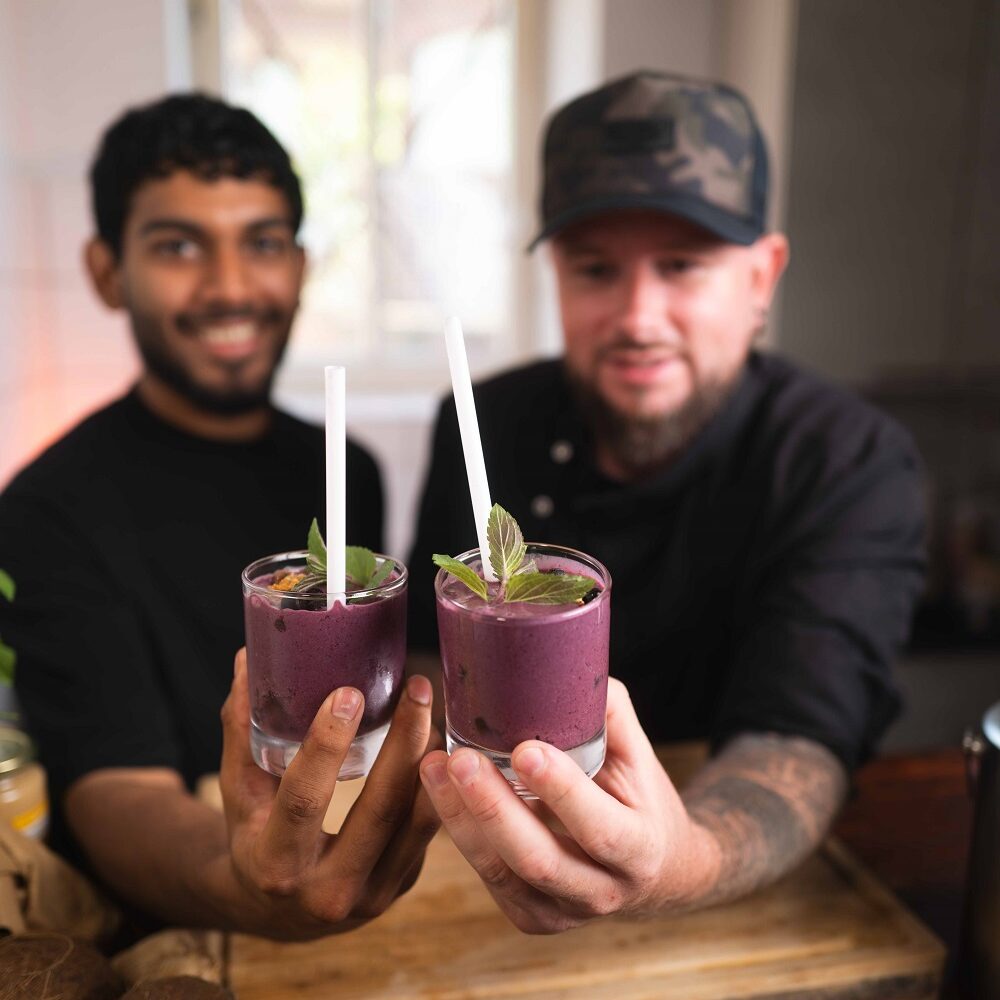 Zwei Männer lächeln und halten lilafarbene Smoothies mit Minzblättern und Strohhalmen in die Höhe. Sie stehen in einer Küche mit einer Holztheke, Kokosnüssen und einem Fenster im Hintergrund.