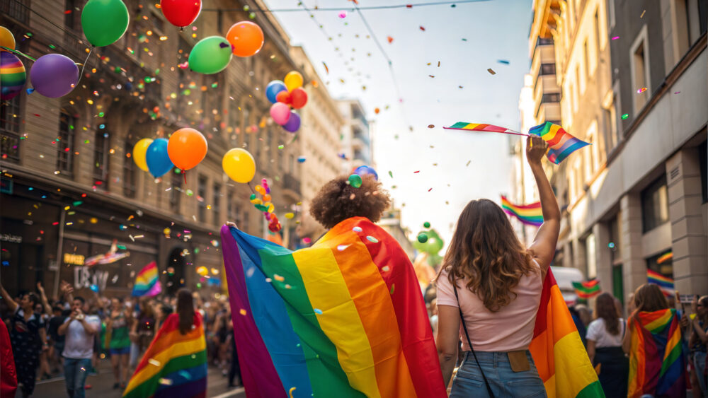 Zwei Menschen laufen durch eine lebhafte Pride-Parade und schwenken Regenbogenflaggen. Bunte Luftballons und Konfetti füllen die Luft über einer von Menschen gesäumten Straße.
