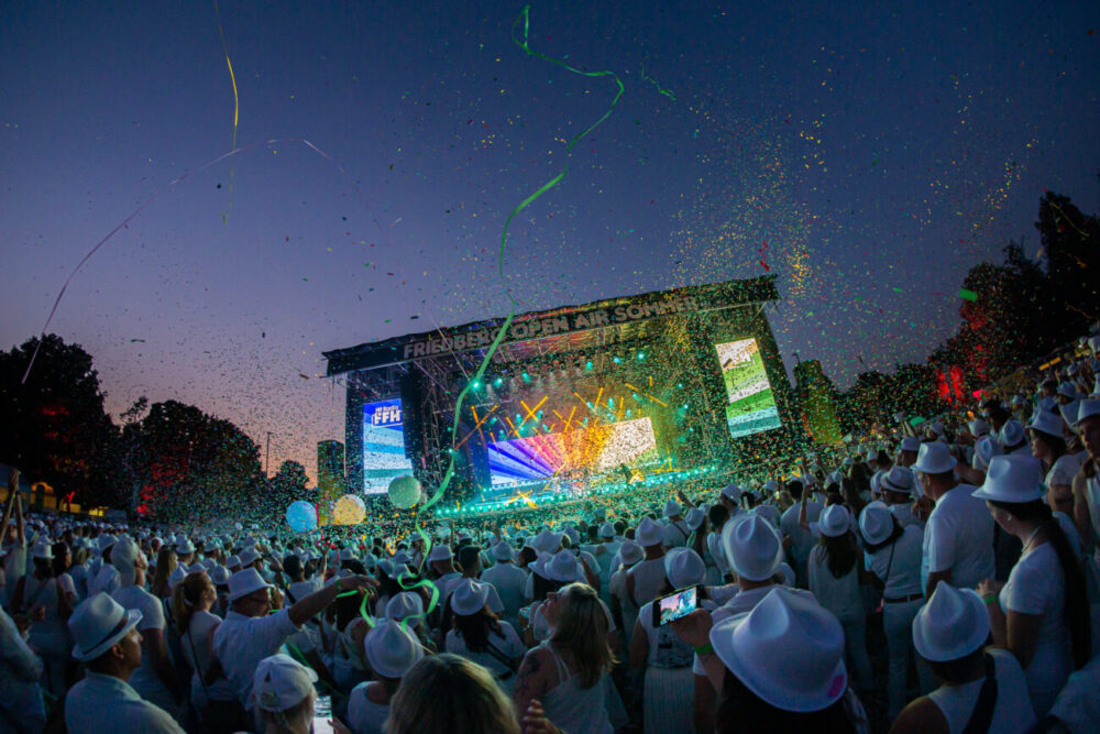 Eine große Menschenmenge in weißen Kleidern und Hüten genießt ein lebhaftes Konzert im Freien in der Abenddämmerung, mit bunten Lichtern auf der Bühne, Konfetti und Luftschlangen in der Luft. Viele Menschen nehmen das Ereignis mit ihren Handys auf.